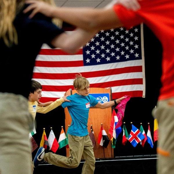 六年级学生围成一圈跳舞，背景是美国国旗，小国旗也在展示