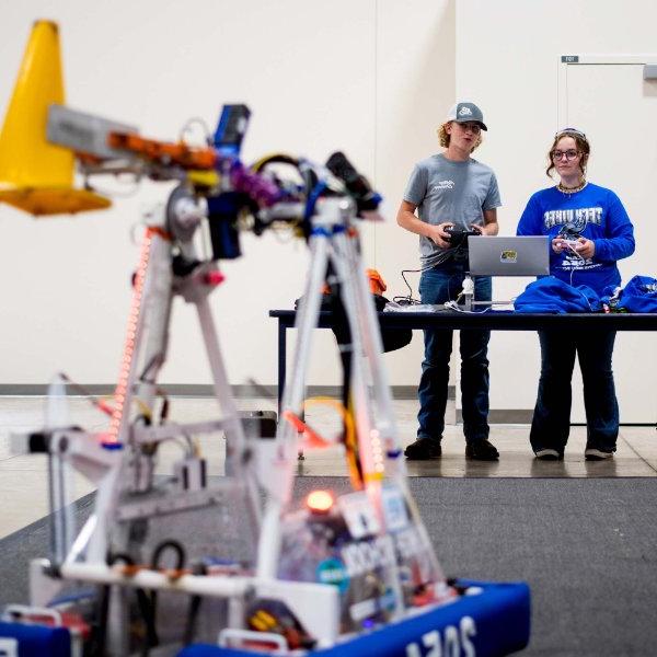 two people stand at a table, a robot from FIRST Robotics in the foreground