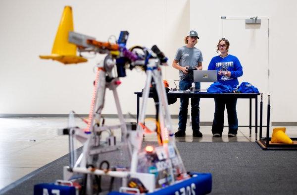 两个人站在桌旁，前景是FIRST机器人公司的机器人