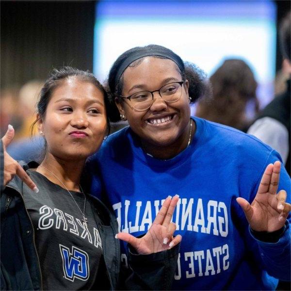 Two students in Grand Valley shirts smile together and hold their fingers and thumb in an Anchor Up pose.